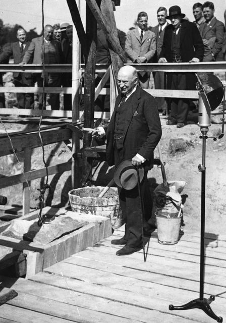 Kuvassa Henri de Baillet-Latour, muurauslasta kädessään, toisessa hattu ja kävelykeppi. Hän seisoo puisella alustalla laastiastioiden vierellä. Taustalla ihmisjoukko katsoo.ö